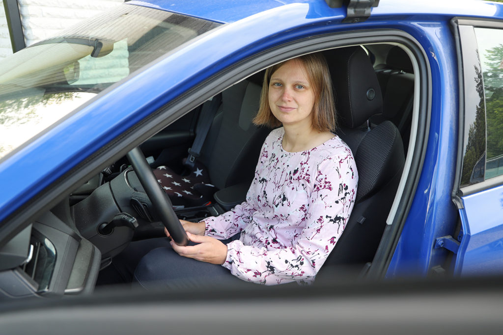 Polkkatukkainen nainen istuu etuovi avoinna olevan sinisen auton ratissa. ja katsoo kameraan.