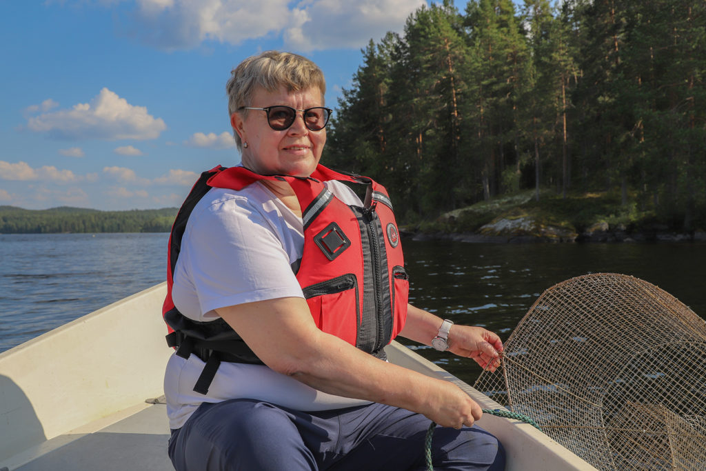 Lyhyttukkainen aurinkolaseihin ja pelastusliiveihin sonnustautunut nainen istuu veneen perässä ja nostaa katiskaa järvestä. 