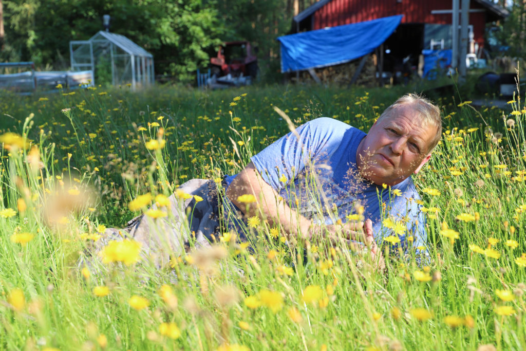 Mies makoilee kyljellään kyynärpäähän nojaten kukkien keskellä korkeassa nurmessa.