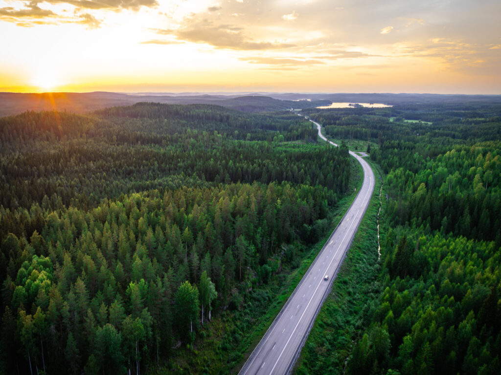 Kuvassa tie, jonka molemmin puolin metsää. Horisontissa on auringonlasku ja taivas on kellertävä.